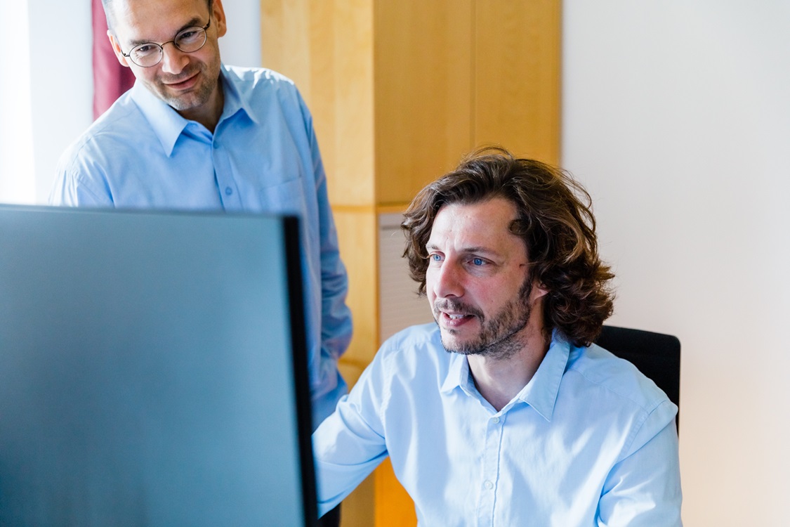 2 Personen, einer sitzend und einer stehend vor einem Computerbildschirm